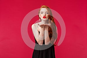 Portrait of coquettish feminine young beautiful redhead woman in black stylish dress, sending air kiss on palms near