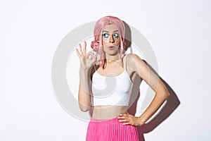 Portrait of coquettish party girl in pink wig and halloween outfit, showing okay sign and looking at upper left corner