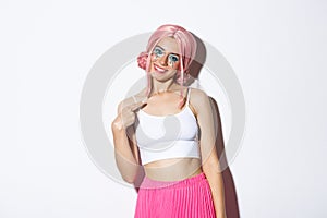 Portrait of coquettish pretty girl in pink wig and halloween costume, pointing at herself with lovely smile, standing