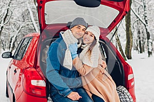 Portrait of couple in love hugging in car trunk in snowy winter forest. People relaxing outdoors during road trip