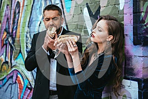 portrait of couple in luxury clothing with hot dogs standing near wall with graffiti