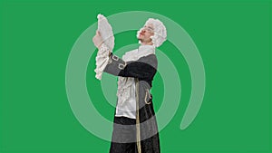 Portrait of courtier gentleman in black historical vintage suit and white wig, waving a fan and curtsy. Young man posing