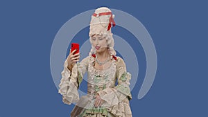 Portrait of courtier lady in white vintage lace dress and wig talking for video chat using mobile phone. Young woman