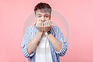 Portrait of coward, nervous scared brown-haired man in casual striped shirt biting finger nails. isolated on pink background