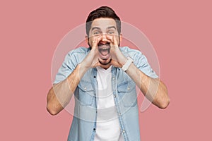 Portrait of crazy handsome bearded young man in blue casual style shirt standing with hand on face, looking at camera and