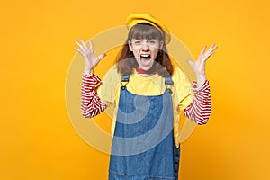Portrait of crazy screaming girl teenager in french beret, denim sundress spreading hands isolated on yellow wall
