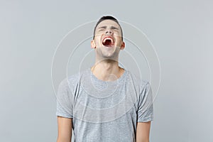 Portrait of crazy screaming young man in casual clothes keeping eyes closed isolated on grey wall background in studio