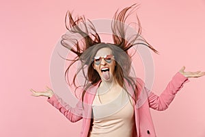 Portrait of crazy young woman in heart glasses showing tongue jumping with flying hair isolated on pastel pink wall