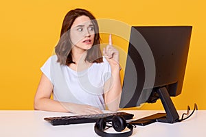 Portrait of creative brunette haired woman, wearing white casual t shirt, sitting in front of computer and pointing finger up, photo