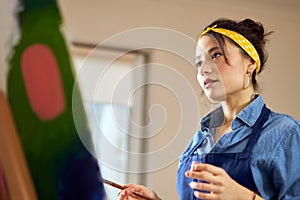 Portrait of creative young woman in apron looking at her work while working on abstract painting at cozy workshop