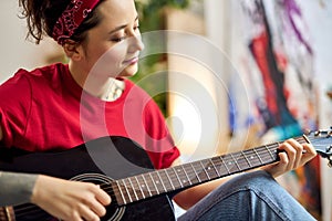 Portrait of creative young woman in casual wear playing acoustic guitar while studying music at home