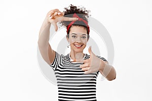 Portrait of creative young woman in headband, picturing moment, looking through hand frames gesture and smiling