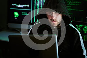 Portrait of criminal hooded hacker sitting at desk and breaking into government or big company data servers in