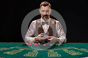 Portrait of a croupier is holding playing cards, gambling chips on table. Black background