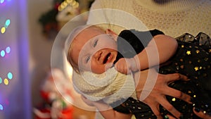 Portrait of crying baby girl in female Caucasian hands at background of Christmas lights. Unsatisfied toddler in