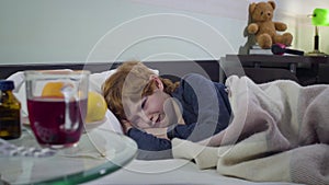 Portrait of crying caucasian little boy lying in bed in front of pills, oranges and hot tea standing on the table. Upset