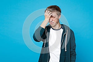 Portrait of crying displeased young man in casual clothes keeping eyes closed putting hand on head isolated on blue