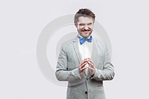 Portrait of cunning handsome bearded man in casual grey suit and blue bow tie standing with funny face and palm hand looking at