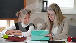 Portrait of curios teenage schoolgirl and intelligent beautiful teacher talking sitting with digital tablet at desk