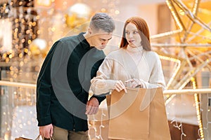 Portrait of curiosity young couple walking shopping mall holding shopping bags, looking inside, excited about their new