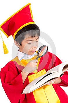 Portrait Of Curious Boy In Red Gown Kid Graduation With Mortarboard Looking A Book Through Magnifying Glass