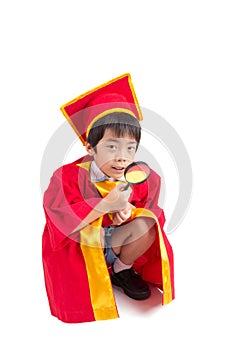 Portrait Of Curious Boy In Red Gown Kid Graduation With Mortarboard Looking Something Through Magnifying Glass