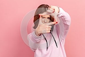 Portrait of curious brunette teenage girl with curly hair in hoodie looking through photo frame gesture made of hands, capturing