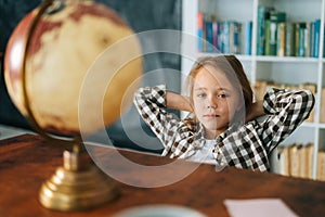Portrait of curious child girl sitting on chair with hands behind head in front of table with a small globe and looking