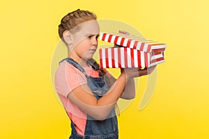 Portrait of curious nosy little girl with braid in denim overalls peeking into gift box, in anticipation of surprise photo