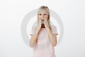 Portrait of curious serious girl with blond hair, holding palms near opened mouth and whispering, telling secret to