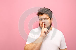 Portrait of a curly overweight boy on a pink background, looks into the camera and shows a gesture of silence with his finger. Fat