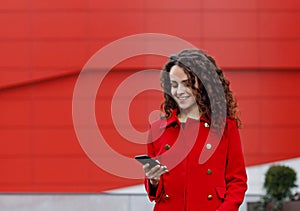 Portrait of a curly young brunette young woman, wear in red jacket, read messages, over building background.