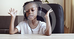 Portrait of cute active African little preschool girl sitting at table at home, wearing headphones talking on headset