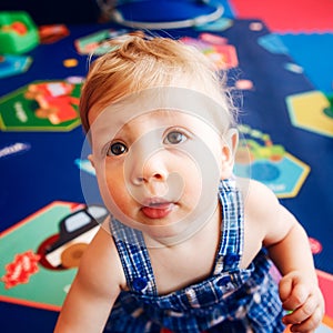 Portrait of cute adorable blonde Caucasian smiling baby boy with brown eyes in blue romper sitting on floor in kids room