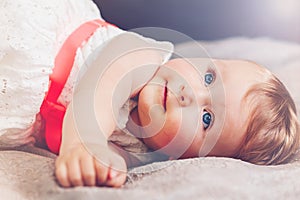 Portrait of cute adorable blonde Caucasian smiling baby child girl with blue eyes in white dress with red bow lying on bed