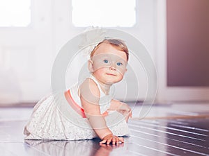 Portrait of cute adorable blonde Caucasian smiling baby child girl with blue eyes in white dress with red bow sitting on floor