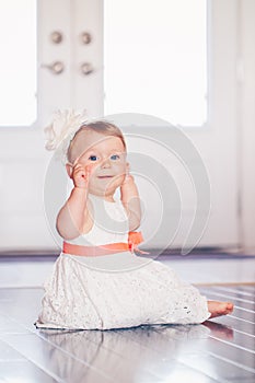 Portrait of cute adorable blonde Caucasian smiling baby child girl with blue eyes in white dress with red bow sitting on floor ind