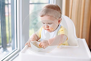 Caucasian child boy with dirty messy face sitting in high chair eating apple puree