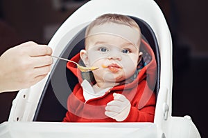 Portrait of cute adorable Caucasian little baby boy sitting in high chair in kitchen eating meal puree