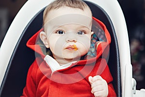 Portrait of cute adorable Caucasian little baby boy sitting in high chair in kitchen eating meal puree