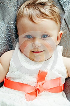 Portrait of cute adorable Caucasian smiling laughing baby girl with blue eyes in white dress with red bow lying on bed looking in