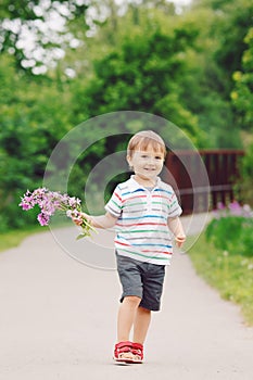 Portrait of a cute adorable funny little smiling boy toddler walking in park with lilac purple pink flowers in hands