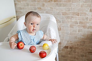 Portrait of cute adorable smiling laughing Caucasian child kid girl sitting in high chair eating apple fruit. Everyday lifestyle.