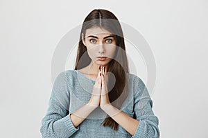 Portrait of cute adult woman with long hair and angel look, glancing at camera while holding hands in pray or beg