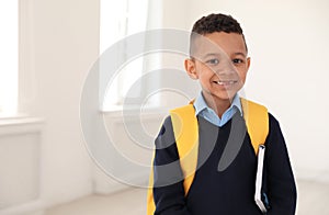 Portrait of cute African-American boy in school uniform with backpack and book indoors