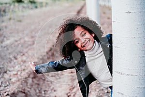 portrait of cute afro kid girl hiding behind a tree outdoors. Childhood and happiness