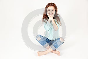Portrait of cute amusing predronee girl with long dark hair in jeans, sitting on the floor and holding her face, isolated on white