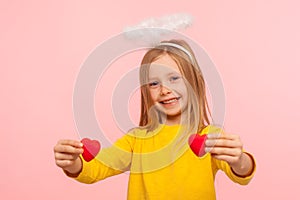 Portrait of cute angelic happy little girl with halo over head holding toy hearts and looking at camera with toothy smile