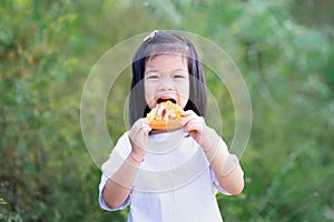 Portrait cute Asian girl is enjoy eating pizza with gusto. Happy child holding food, taking it into her mouth with bite to eat.