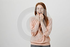 Portrait of cute attractive caucasian ginger girl covering mouth while laughing and expressing excitement, standing